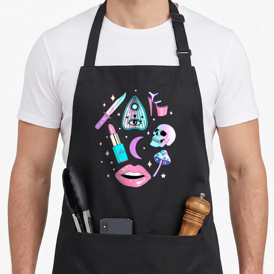 Witch goth apron - Cartoon collage of pink, blue, and purple occult objects including skull, lips, and stars — shown on a black cotton canvas apron against a white background