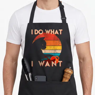 Cat apron for the grill master - I DO WHAT I WANT text around cat silhouette and toy against striped retro sun — shown on a black cotton canvas apron against a white background
