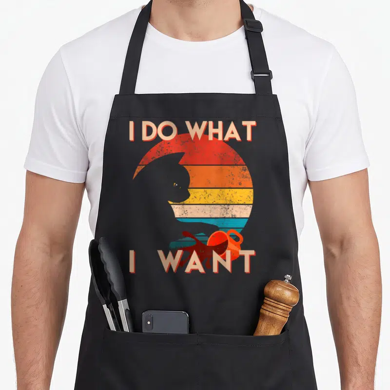 Cat apron for the grill master - I DO WHAT I WANT text around cat silhouette and toy against striped retro sun — shown on a black cotton canvas apron against a white background