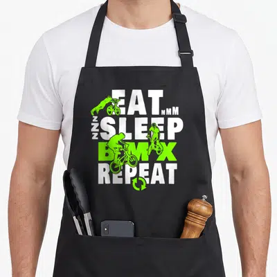 Sleep bicycle apron - Stacked EAT SLEEP BMX REPEAT text with lime green BMX rider silhouettes and a circular repeat — shown on a black cotton canvas apron against a white background