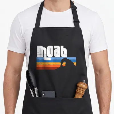Moab arches apron - White moab text over blue orange red stripes with grey mountain silhouette — shown on a black cotton canvas apron against a white background