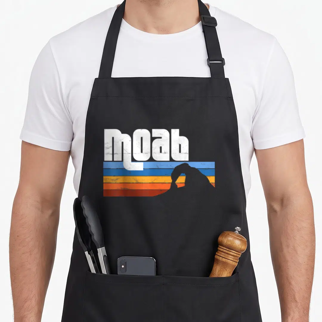 Moab arches apron - White moab text over blue orange red stripes with grey mountain silhouette — shown on a black cotton canvas apron against a white background