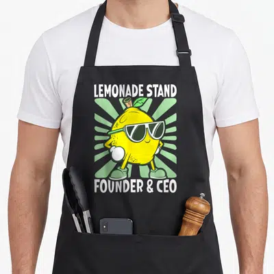 Stand founder lemonade apron - Cartoon yellow lemon wearing sunglasses with green sunburst and white text reading — shown on a black cotton canvas apron against a white background