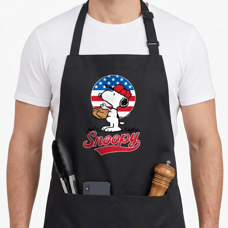 Americana baseball apron - Cartoon dog with brown glove in front of circular red white blue flag and red Snoopy text — shown on a black cotton canvas apron against a white background