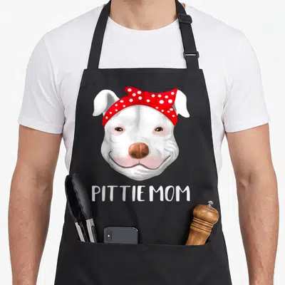Pitbull mother's day aprons - Cartoon white pit bull head with red polka dot bow and PITTIE MOM text below — shown on a black cotton canvas apron against a white background