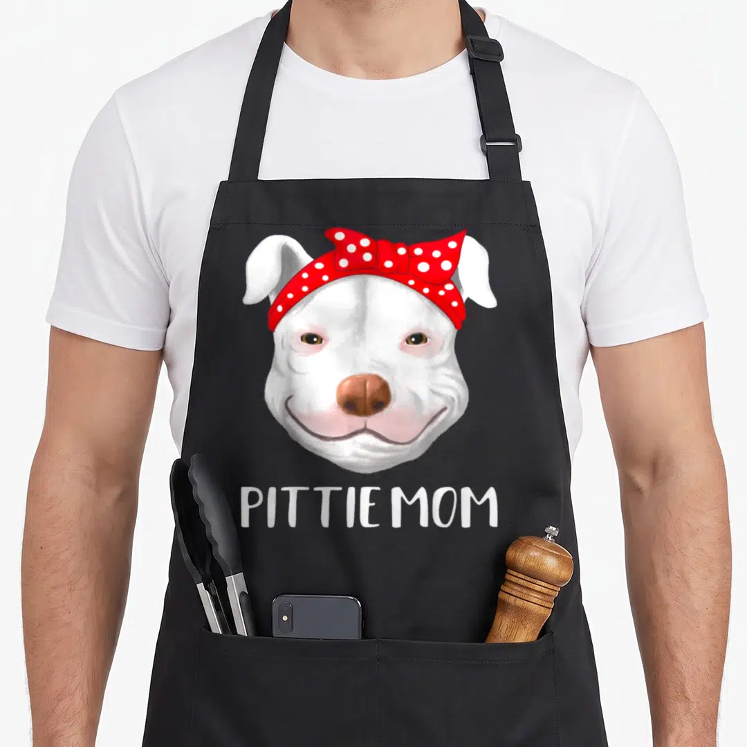 Pitbull mother's day aprons - Cartoon white pit bull head with red polka dot bow and PITTIE MOM text below — shown on a black cotton canvas apron against a white background