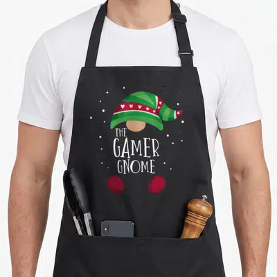 Pjs christmas gnome apron - Cartoon gnome with striped green hat, white text, and two red shoes surrounded by stars — shown on a black cotton canvas apron against a white background