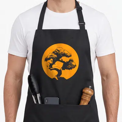 Japanese bonsai tree apron - Grey bonsai tree silhouette inside a solid orange circle on a plain background — shown on a black cotton canvas apron against a white background