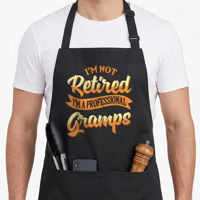 Professional gramp father day apron - Retro orange and yellow text reading I'm Not Retired I'm A Professional Gramps — shown on a black cotton canvas apron against a white background
