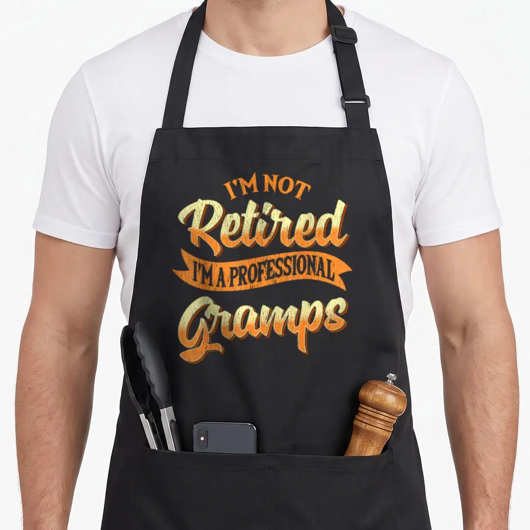 Professional gramp father day apron - Retro orange and yellow text reading I'm Not Retired I'm A Professional Gramps — shown on a black cotton canvas apron against a white background