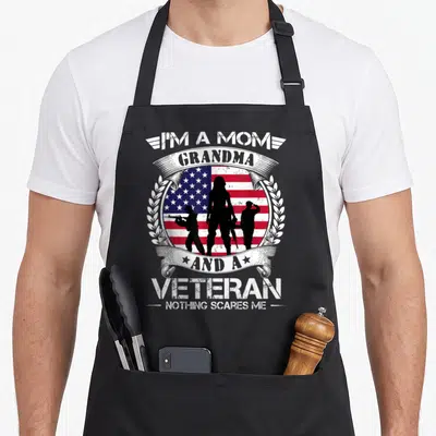 Scare military grandma apron - I'm a mom grandma veteran nothing scares me text with three soldiers and flag — shown on a black cotton canvas apron against a white background