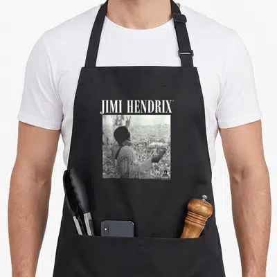Jimi woodstock era apron - JIMI HENDRIX text above square B&W photo of musician with raised hand facing crowd — shown on a black cotton canvas apron against a white background