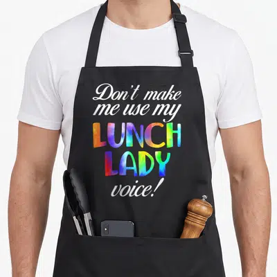 Use lunch lady apron - Stacked text reading Don't make me use my LUNCH LADY voice in tie-dye block letters and white — shown on a black cotton canvas apron against a white background