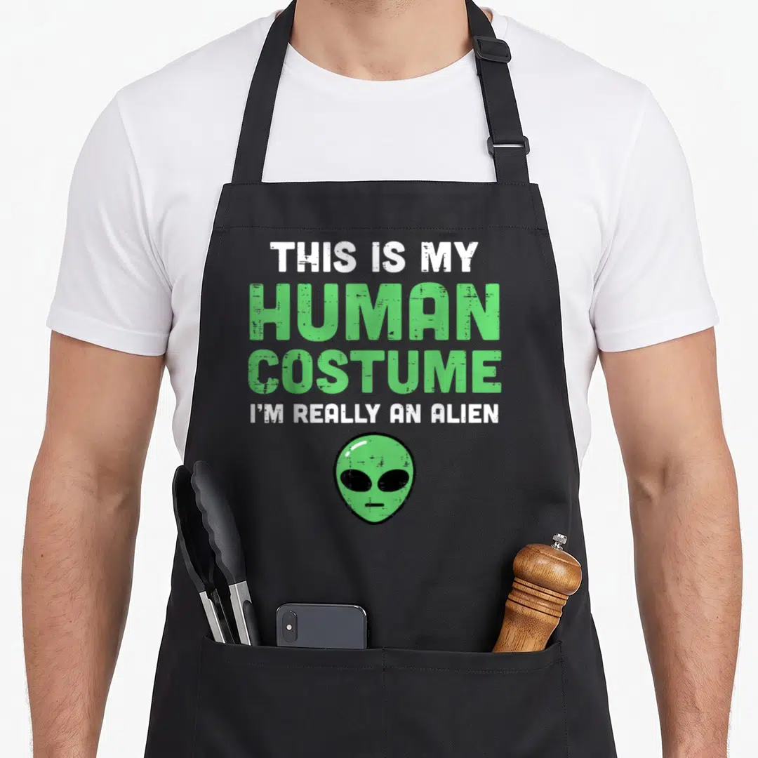 Human costume alien halloween apron - Stacked green and white text above a green cartoon alien head on a black — shown on a black cotton canvas apron against a white background