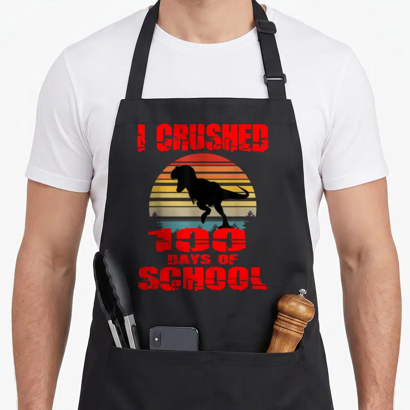 100th dinosaur apron - Red I CRUSHED 100 DAYS OF SCHOOL text over black T-Rex and striped sun — shown on a black cotton canvas apron against a white background