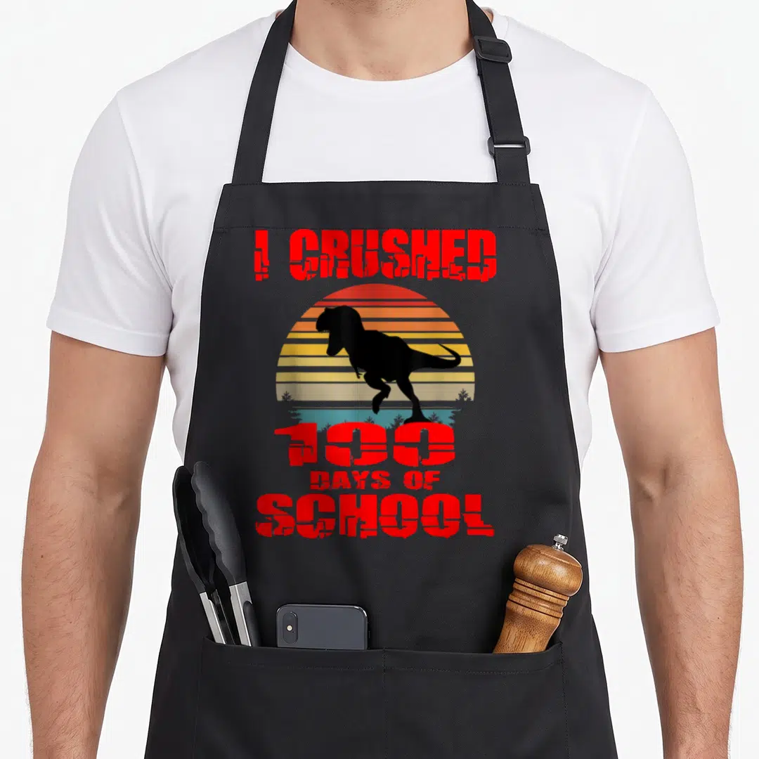 100th dinosaur apron - Red I CRUSHED 100 DAYS OF SCHOOL text over black T-Rex and striped sun — shown on a black cotton canvas apron against a white background