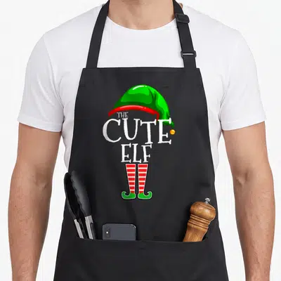 Funny family christmas elf apron - Cartoon elf hat and legs with stacked THE CUTE ELF text in green red white yellow — shown on a black cotton canvas apron against a white background