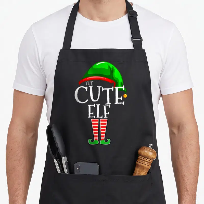 Funny family christmas elf apron - Cartoon elf hat and legs with stacked THE CUTE ELF text in green red white yellow — shown on a black cotton canvas apron against a white background