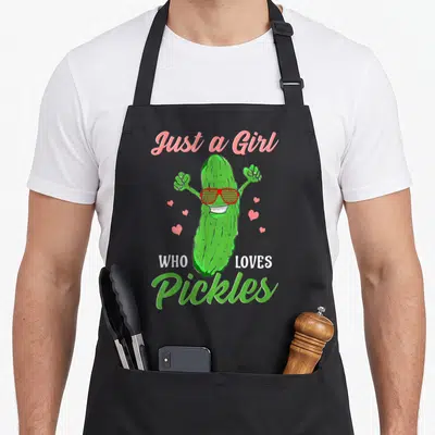 Food pickle apron - Green cartoon pickle wearing red sunglasses with pink hearts and text Just a Girl Who Loves Pickles — shown on a black cotton canvas apron against a white background