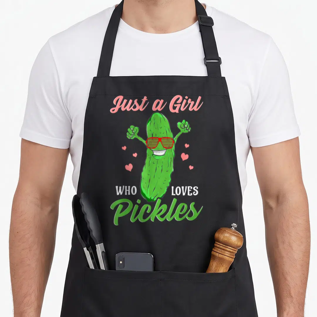 Food pickle apron - Green cartoon pickle wearing red sunglasses with pink hearts and text Just a Girl Who Loves Pickles — shown on a black cotton canvas apron against a white background