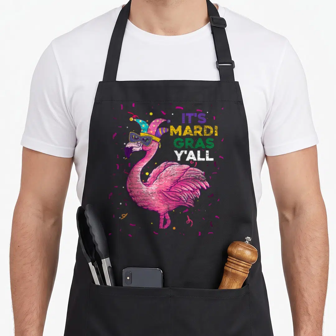Mardi flamingo apron - Pink flamingo in jester hat with It's Mardi Gras Y'all text and confetti — shown on a black cotton canvas apron against a white background