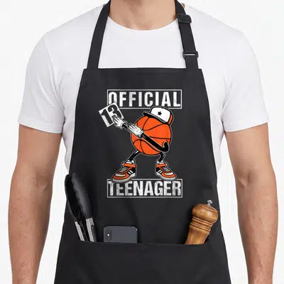 13th basketball birthday apron - Dabbing orange basketball character wearing cap and sneakers with OFFICIAL 13 TEENAGER — shown on a black cotton canvas apron against a white background