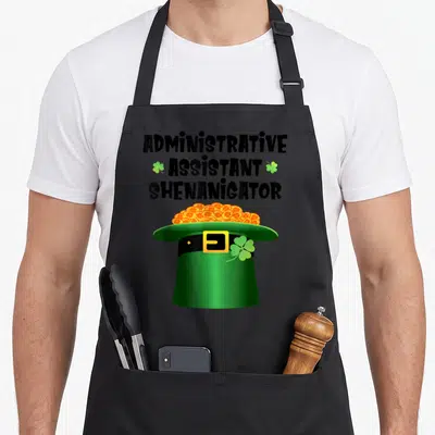 Administrative st patrick day aprons - Administrative Assistant Shenanigator text above green hat with orange coins — shown on a black cotton canvas apron against a white background
