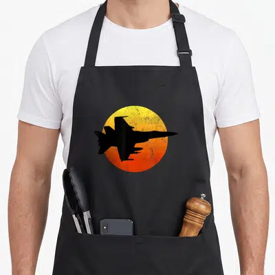 Hornet flying apron - Black fighter jet silhouette over distressed yellow and orange sun circle — shown on a black cotton canvas apron against a white background
