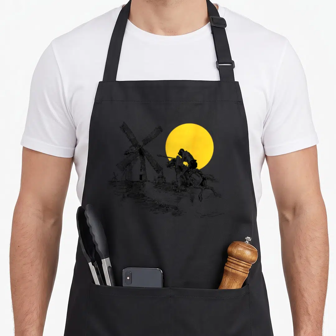 Quixote crazy but apron - Black and white knight on horseback charging a windmill beneath a large yellow circle sun — shown on a black cotton canvas apron against a white background