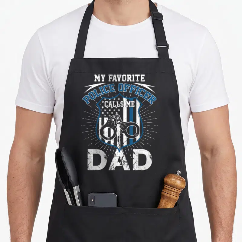 Police officer father day apron - Distressed blue and white badge with handcuffs and flag stripes and My Favorite — shown on a black cotton canvas apron against a white background