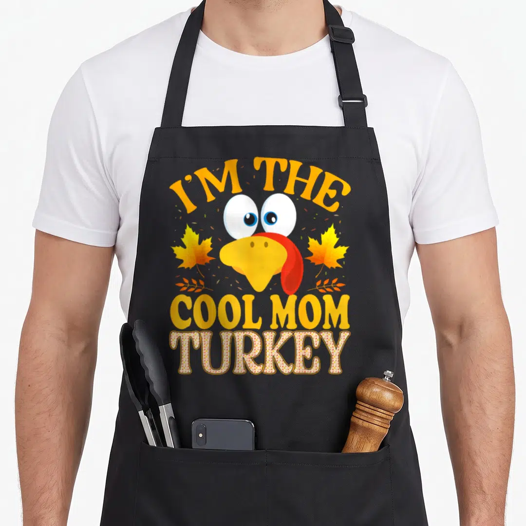 Cute thanksgiving funny turkey apron - Cartoon turkey face with I'M THE COOL MOM TURKEY text in yellow and orange — shown on a black cotton canvas apron against a white background
