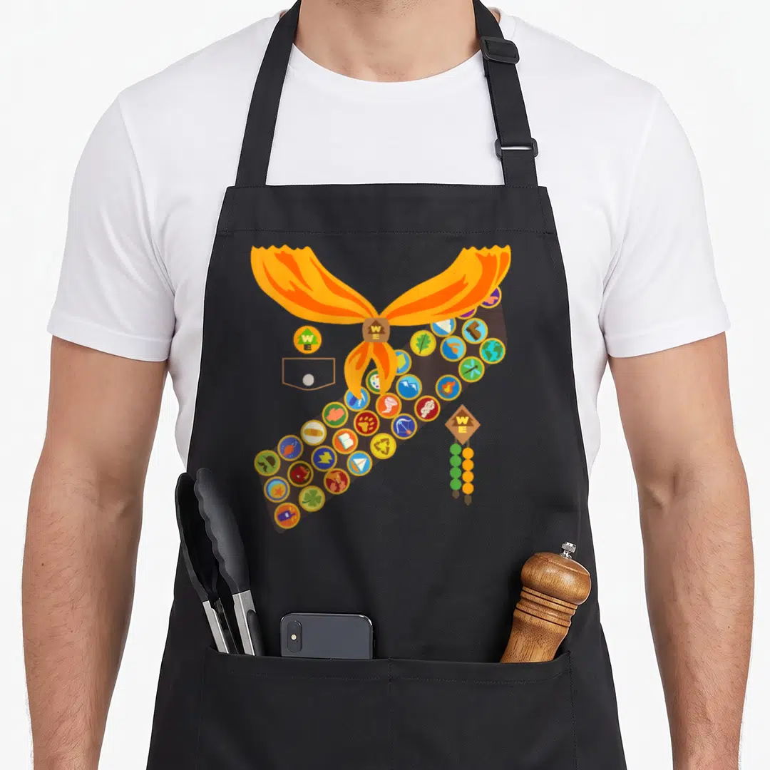 Russell halloween white apron - Cartoon brown sash with circular badges and orange neckerchief tied at top — shown on a black cotton canvas apron against a white background