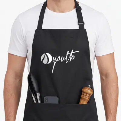 Ignite flame apron - white circular flame logo next to script youth text on canvas — shown on a black cotton canvas apron against a white background