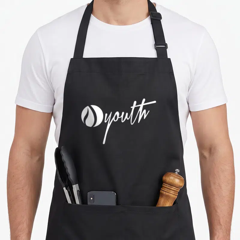 Ignite flame apron - white circular flame logo next to script youth text on canvas — shown on a black cotton canvas apron against a white background