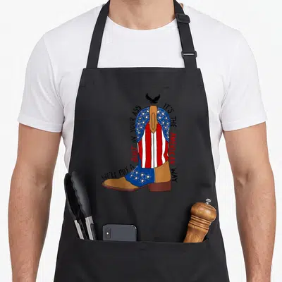 Well western apron - Cartoon cowboy boot with flag pattern and We'll Put A Boot In Your Ass text — shown on a black cotton canvas apron against a white background