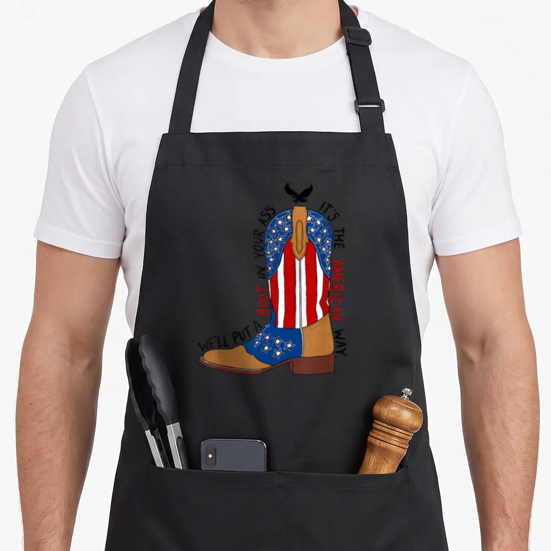 Well western apron - Cartoon cowboy boot with flag pattern and We'll Put A Boot In Your Ass text — shown on a black cotton canvas apron against a white background