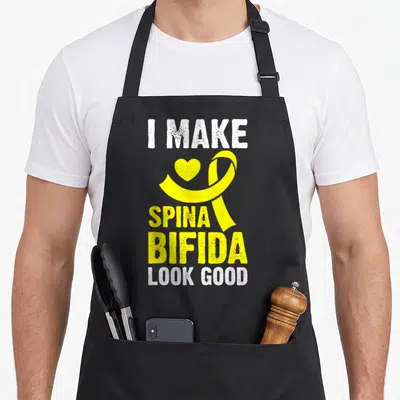 Occulta spina bifida awareness apron - Yellow ribbon and heart between I MAKE SPINA BIFIDA LOOK GOOD white and yellow — shown on a black cotton canvas apron against a white background