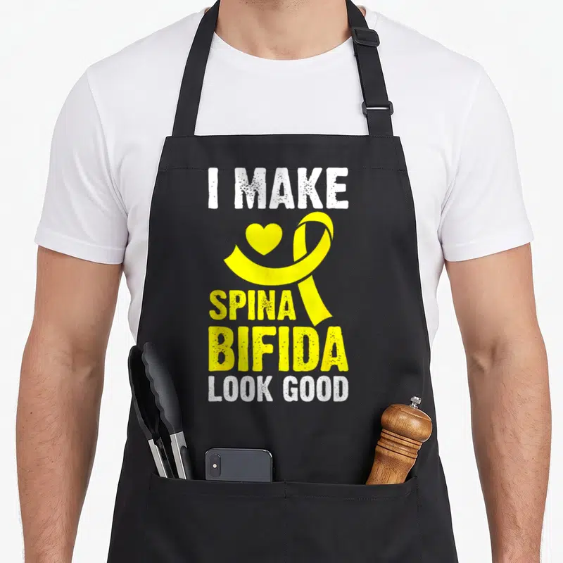 Occulta spina bifida awareness apron - Yellow ribbon and heart between I MAKE SPINA BIFIDA LOOK GOOD white and yellow — shown on a black cotton canvas apron against a white background