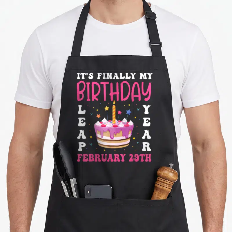 Finally leap birthday apron - Cartoon cake with IT'S FINALLY MY BIRTHDAY text and February 29th date — shown on a black cotton canvas apron against a white background