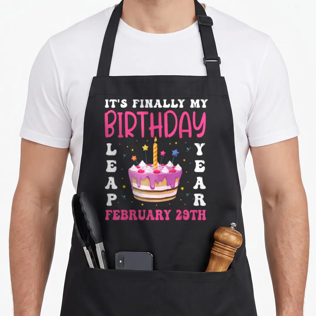 Finally leap birthday apron - Cartoon cake with IT'S FINALLY MY BIRTHDAY text and February 29th date — shown on a black cotton canvas apron against a white background