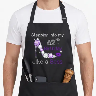 62nd bbq boss apron - Purple high heel shoe and rhinestone text Stepping into my 62ND Birthday Like a Boss — shown on a black cotton canvas apron against a white background