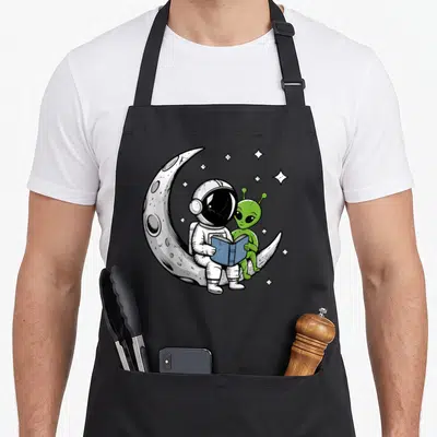 Reading astronaut space apron - Cartoon astronaut and green alien reading blue book on white crescent moon with stars — shown on a black cotton canvas apron against a white background