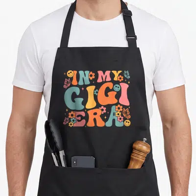 Era mother's day aprons - In My Gigi Era text with smiley faces and flowers in pink blue yellow orange — shown on a black cotton canvas apron against a white background