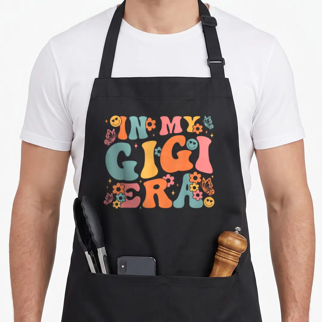 Era mother's day aprons - In My Gigi Era text with smiley faces and flowers in pink blue yellow orange — shown on a black cotton canvas apron against a white background