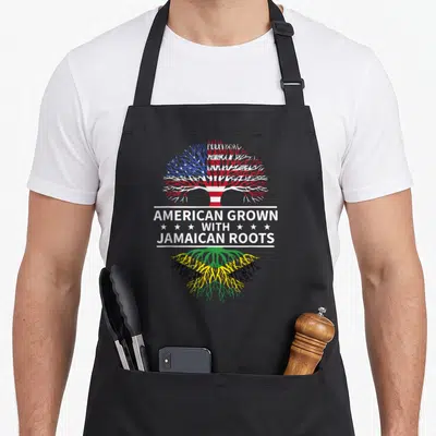 Grown creole apron - Tree with US flag canopy and Jamaica flag roots split by white banner with text — shown on a black cotton canvas apron against a white background