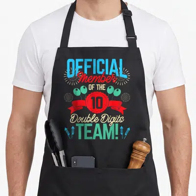 Digit twin birthday apron - Official Member of the 10 Double Digits Team text with red ribbon, balloons, and fireworks — shown on a black cotton canvas apron against a white background