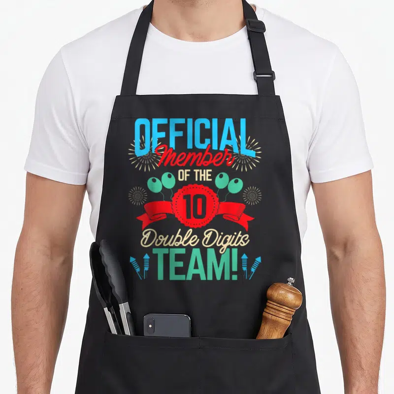 Digit twin birthday apron - Official Member of the 10 Double Digits Team text with red ribbon, balloons, and fireworks — shown on a black cotton canvas apron against a white background