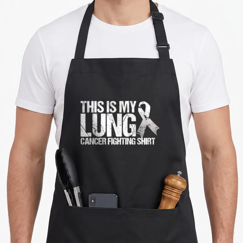 Lung cancer fighter apron - Distressed white text reading THIS IS MY LUNG CANCER FIGHTING SHIRT with ribbon — shown on a black cotton canvas apron against a white background