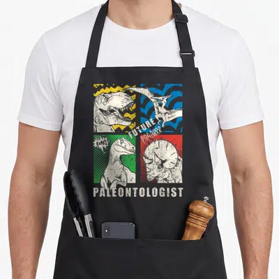 Explorer dinosaur apron - Retro four-color quadrant print with dinosaur sketches and Future Paleontologist text — shown on a black cotton canvas apron against a white background