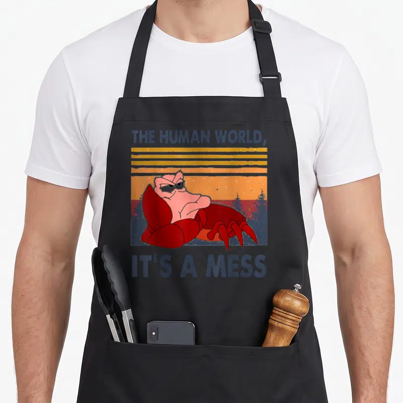 Human crab apron - Red crab with crossed claws under retro sunset and text The Human World It's A Mess — shown on a black cotton canvas apron against a white background